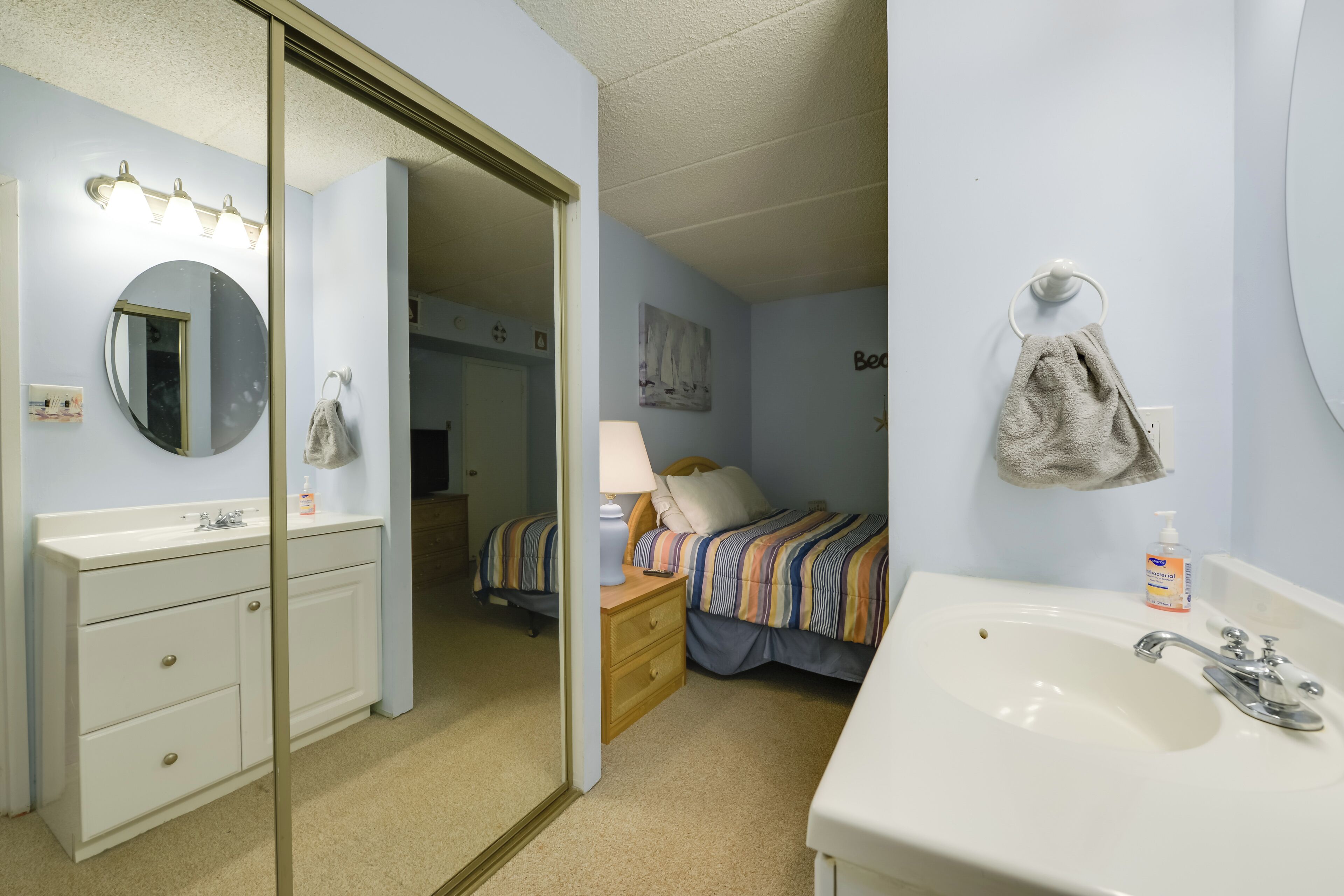 Un dormitorio con un lavamanos blanco, un espejo, una mesita de noche con una lámpara, una cama con un edredón a rayas y una toalla colgada en la pared.