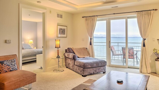 A hotel room with a bed, a sofa, a dining table, and a view of the sea through large windows. - Beachfront Traverse City Resort Condo w/ Pool (Traverse City)