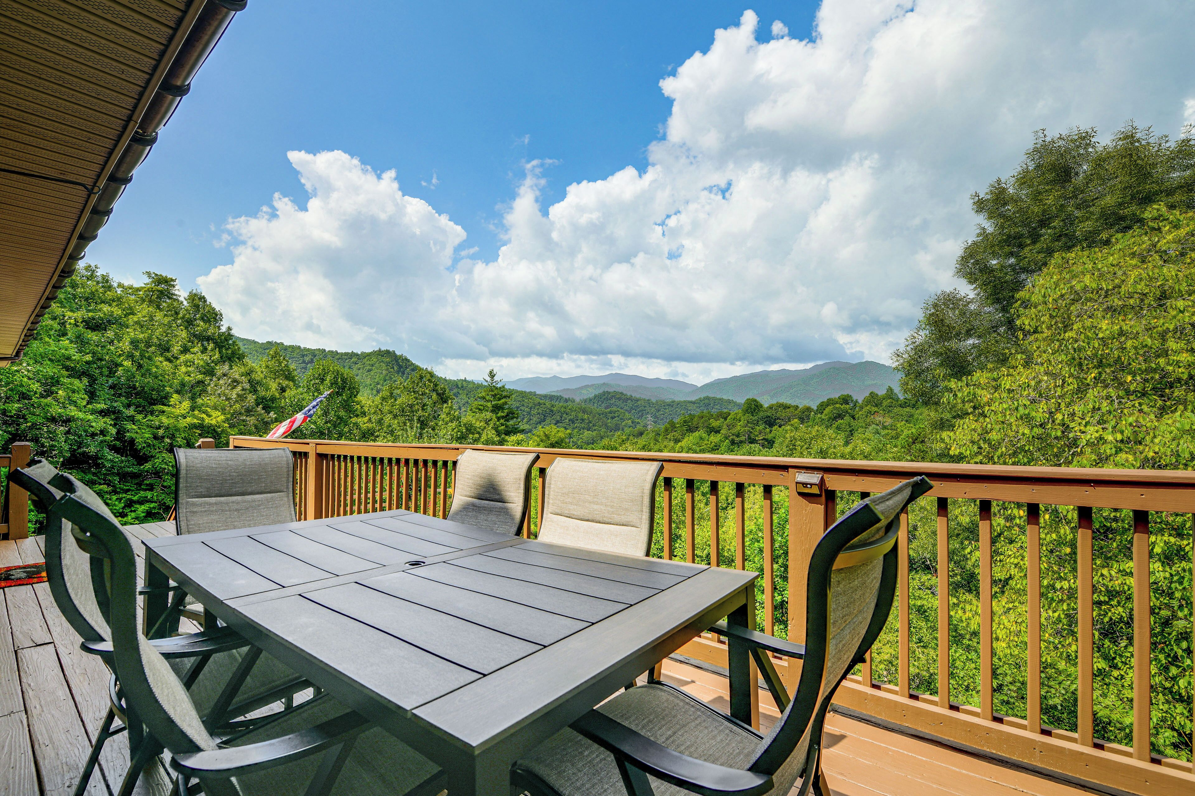 A deck with a table and chairs, overlooking a forested landscape with mountains in the distance.