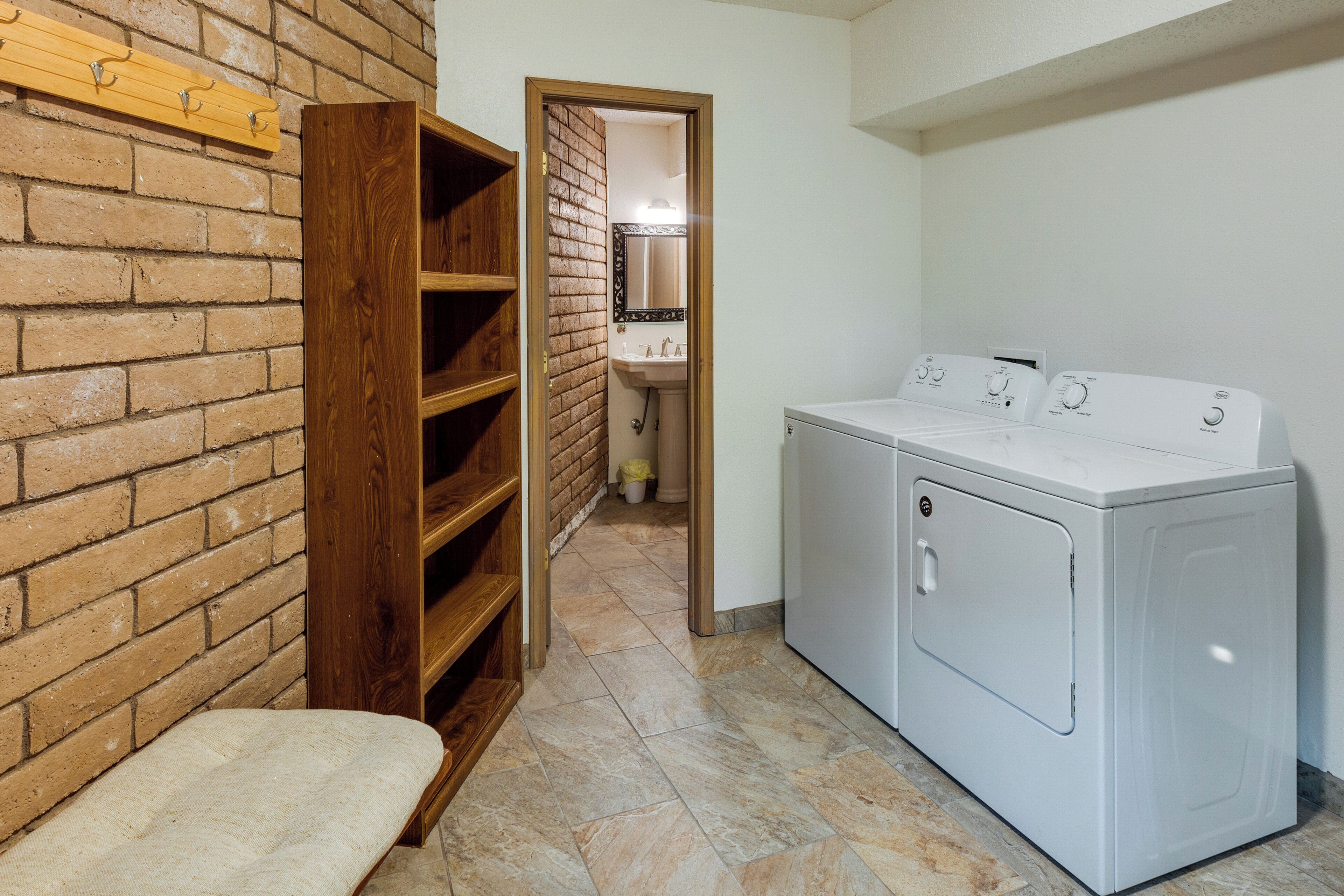Une buanderie avec un mur en briques, des étagères en bois, un banc et un lave-linge blanc.