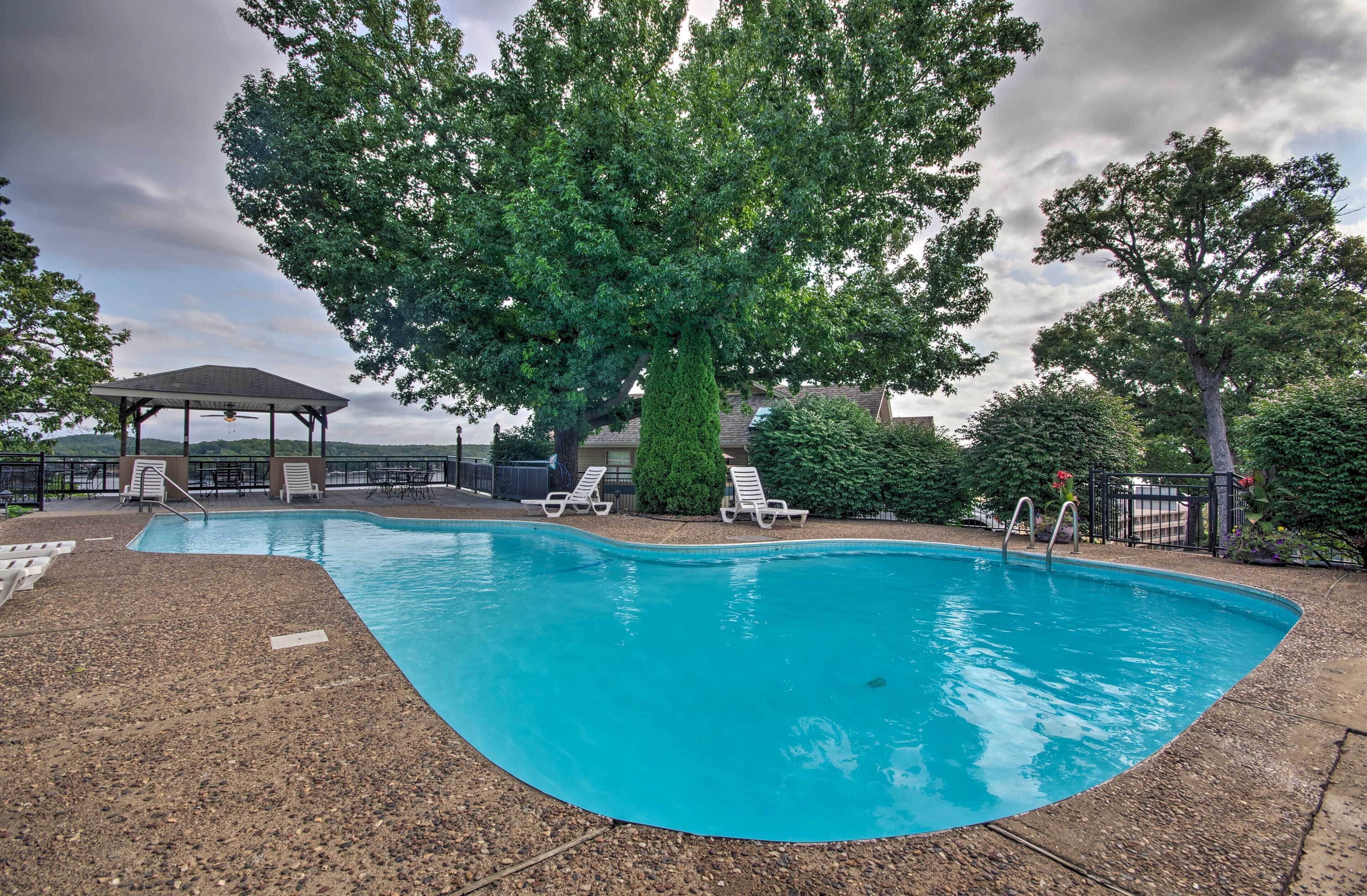 A swimming pool with lounge chairs and a covered seating area, surrounded by trees and a cloudy sky.