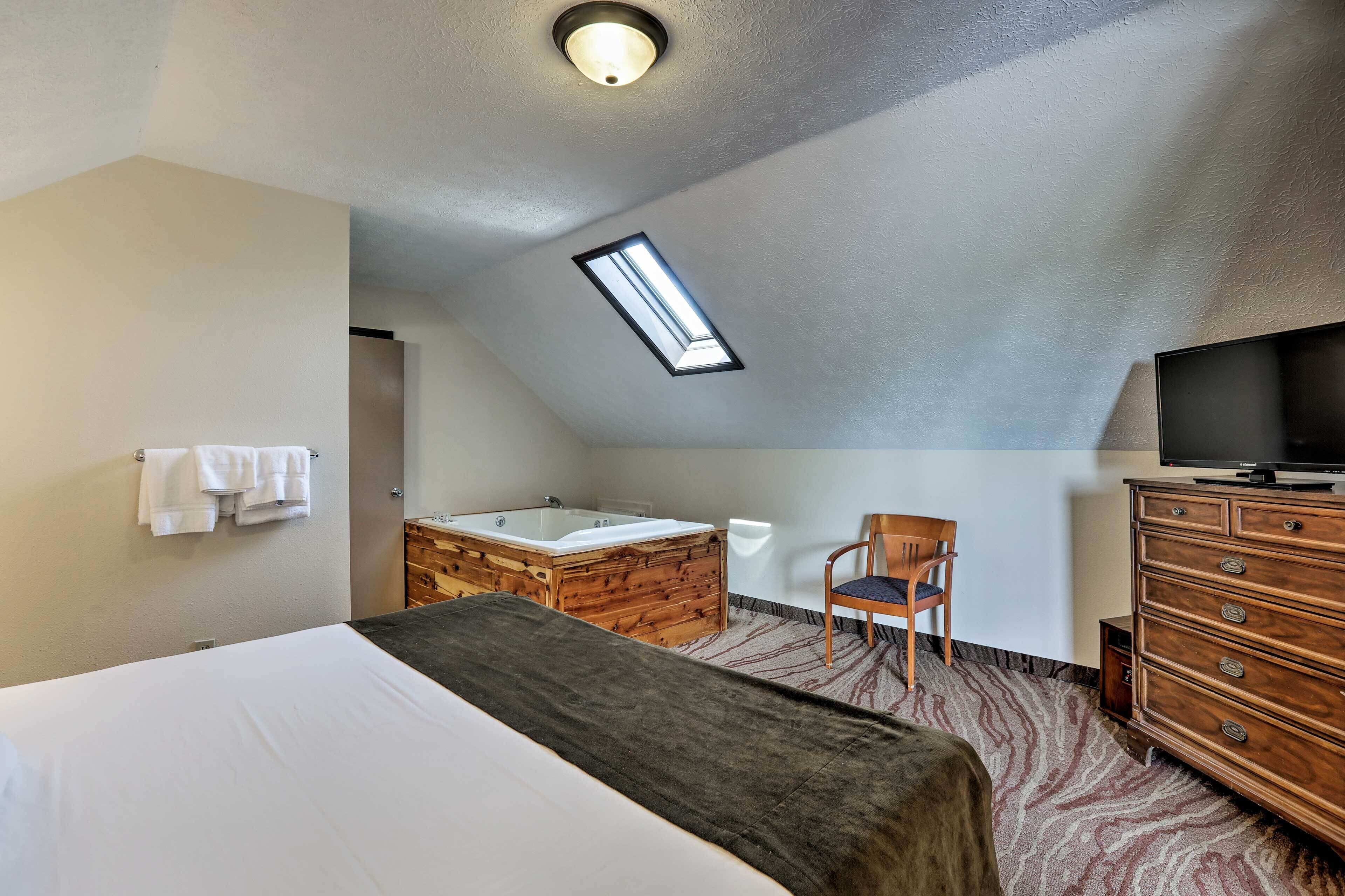 Ein Schlafzimmer mit einem großen Bett, einer hölzernen Kommode, einem Fernseher, einem Sessel und einem Badezimmer mit Badewanne und Dachfenster.