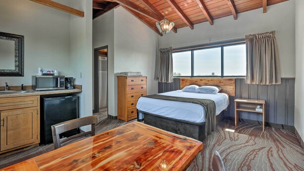 A compact hotel room with a wooden ceiling, a bed with a headboard, a small desk with a microwave, a wooden table, and a window with curtains. - Burkesville Apt w/ Deck, Views & Pool Access! (Burkesville)