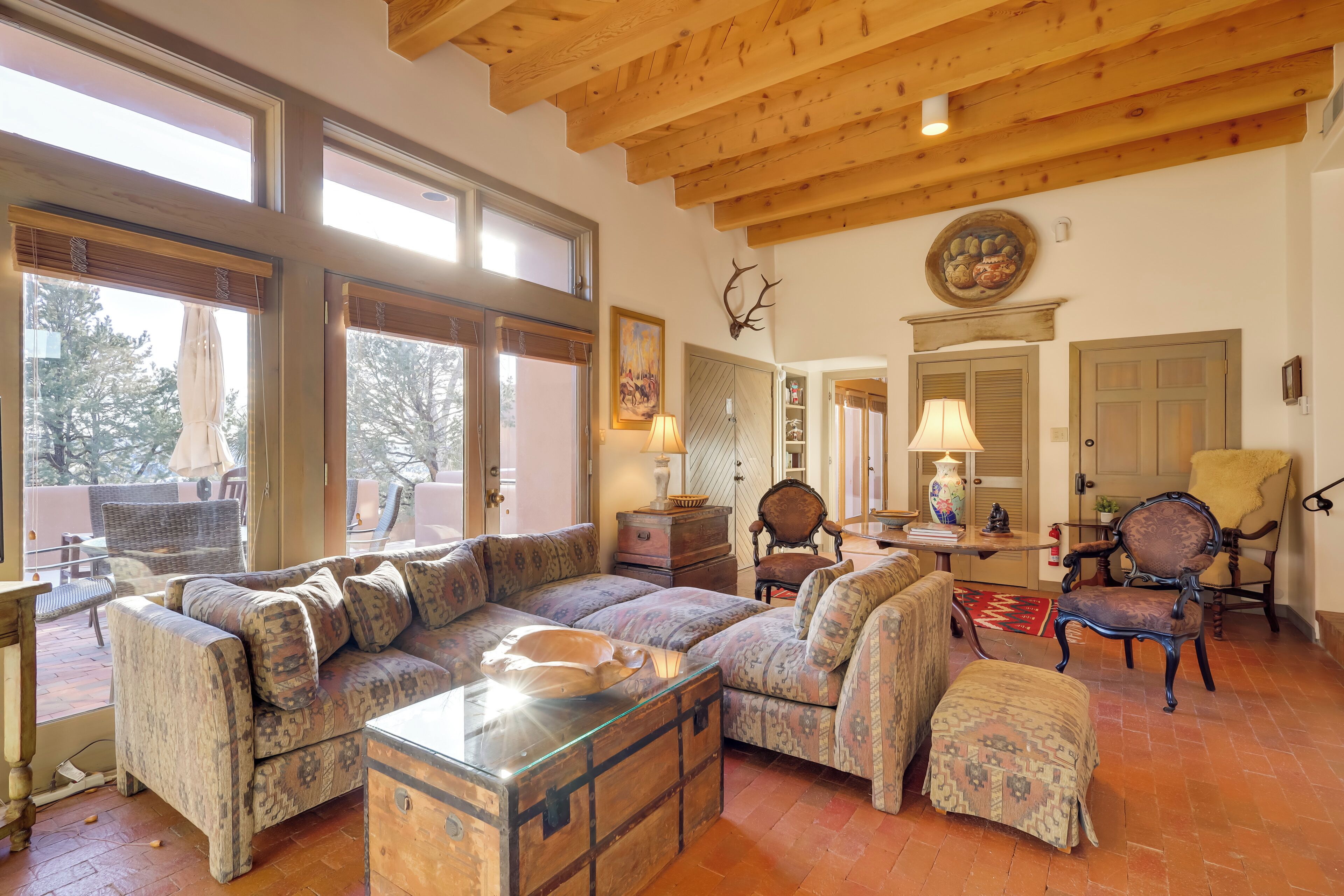A cozy living room with a wooden ceiling, a large sofa, a patterned ottoman, and a rustic chest of drawers.