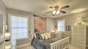 Une chambre à coucher comprenant un lit, un ventilateur de plafond, une fenêtre avec des stores, une table de chevet avec une lampe, une commode et un placard.