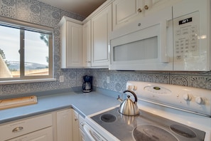 A kitchen with white cabinets, a microwave, a kettle, and a window with a view of the outdoors.