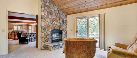 A cozy living room with a stone fireplace, wooden ceiling, and a view through a large window.