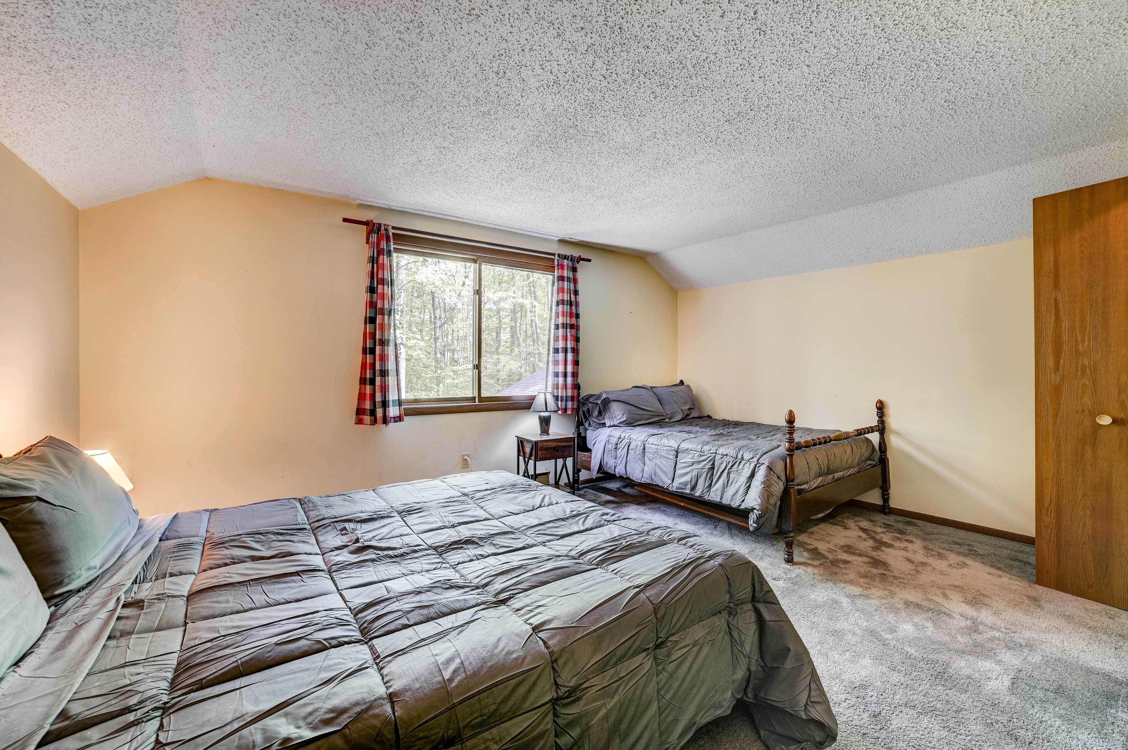 A bedroom with two beds, a window with plaid curtains, a bedside table with a lamp, and a door.