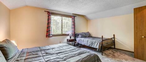 A bedroom with two beds, a window with plaid curtains, a bedside table with a lamp, and a door.