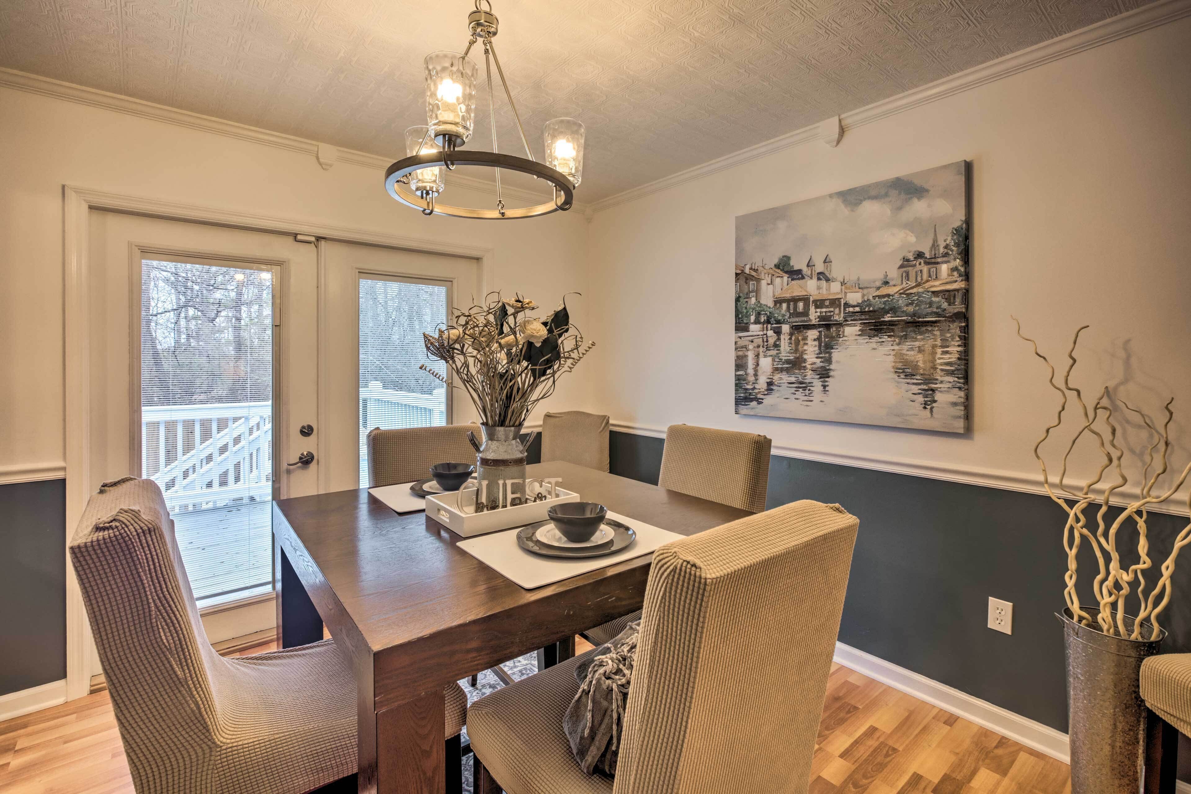 Un coin repas comprenant une table en bois, six chaises, un lustre, un vase avec des fleurs et un tableau accroché au mur.