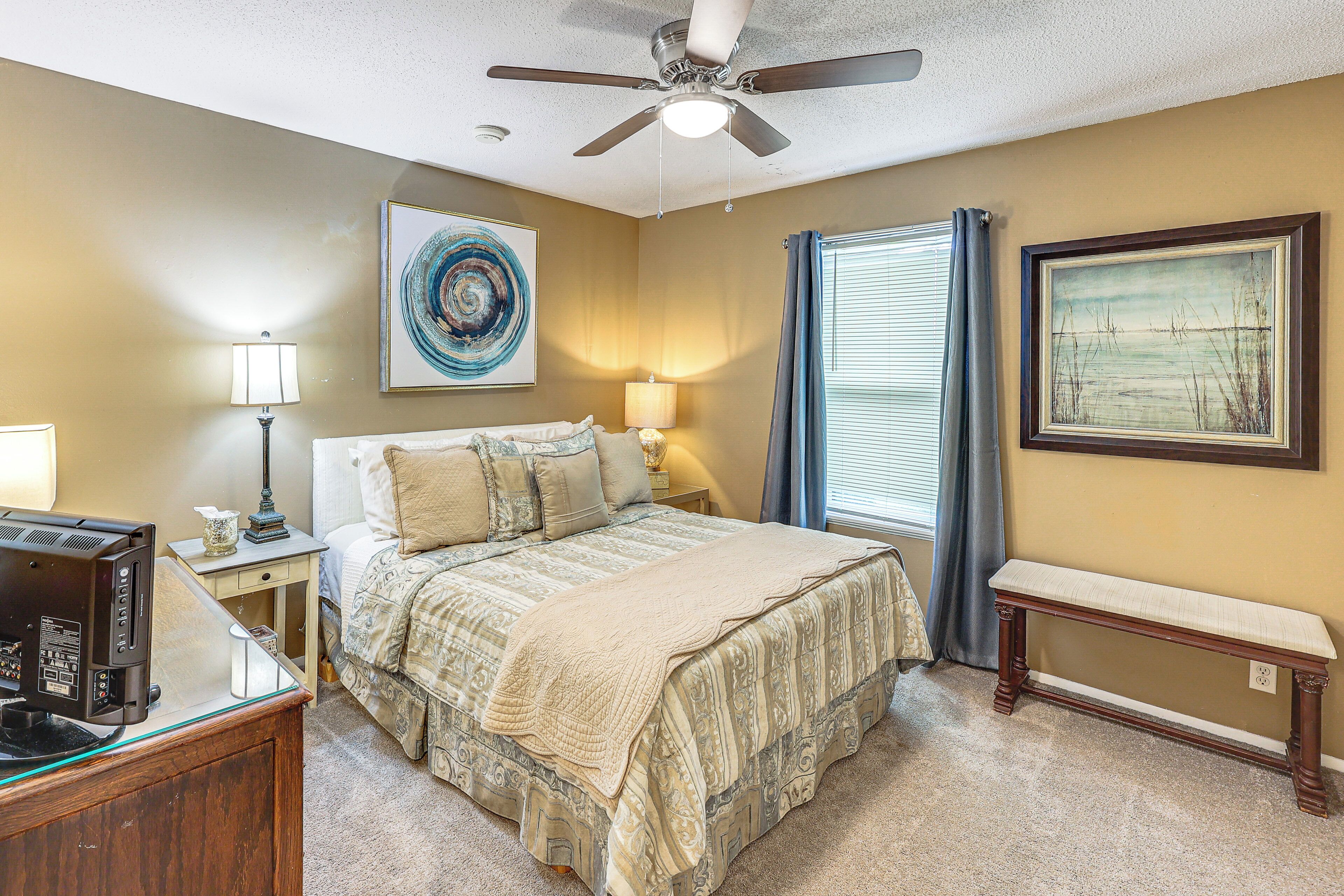 A bedroom with a bed, bedside lamps, a ceiling fan, a desk with a computer, and artwork on the walls.