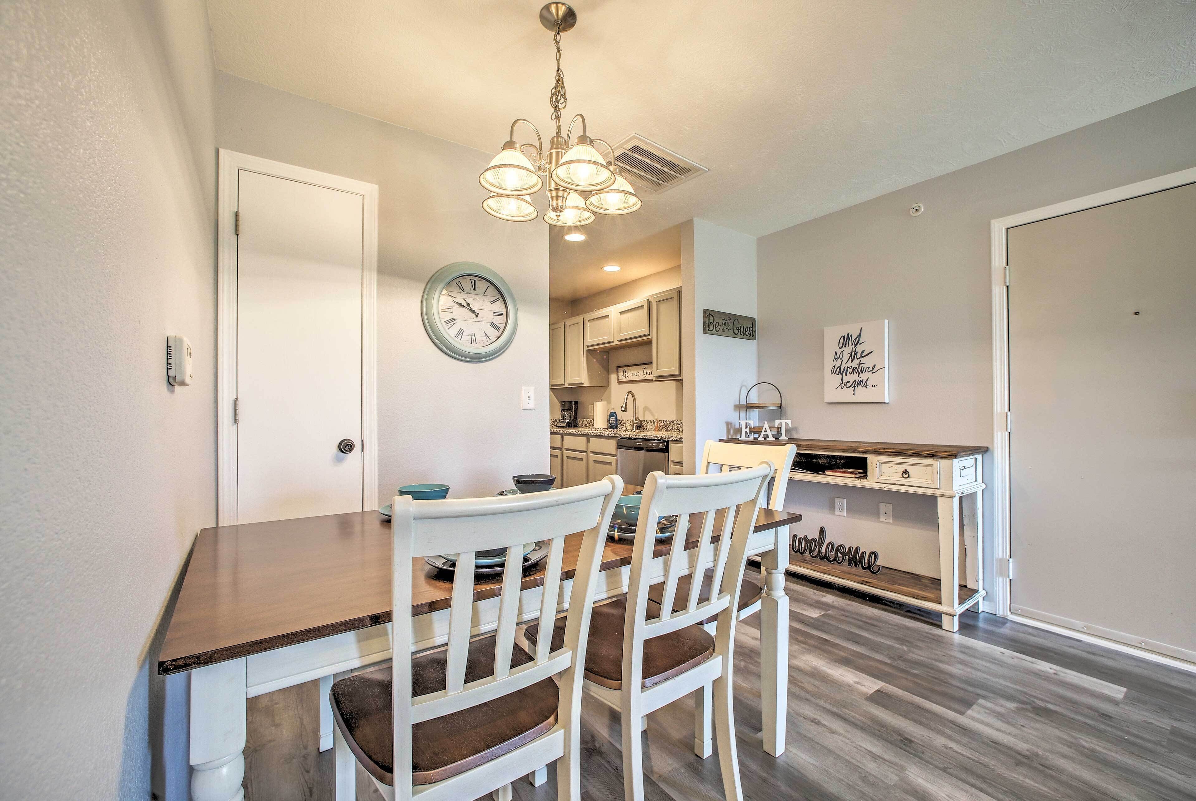 A dining area with a wooden table and white chairs, a clock on the wall, and a kitchen in the background.