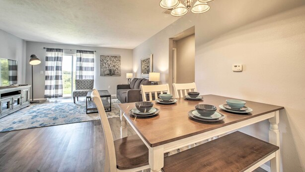 A dining area with a wooden table set for four, surrounded by chairs. A chandelier hangs above the table, and there is a view of a living room with a sofa and a lamp in the background. - Cozy Condo w/ Pool Access in the Heart of Branson! (Branson)