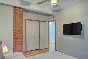 A bedroom with a brick accent wall, a ceiling fan, a bed, and a TV mounted on the wall.