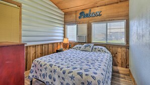 Un dormitorio con cama, paredes de madera, una ventana con persianas y una mesita de noche con lámpara, además de un letrero que dice “Relax”.