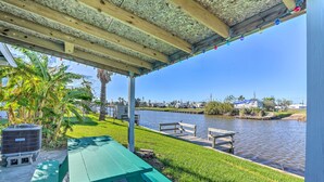 Un espacio al aire libre cubierto con una mesa de picnic verde, un muelle que se extiende sobre un cuerpo de agua tranquilo y una vista despejada de una zona residencial al fondo.