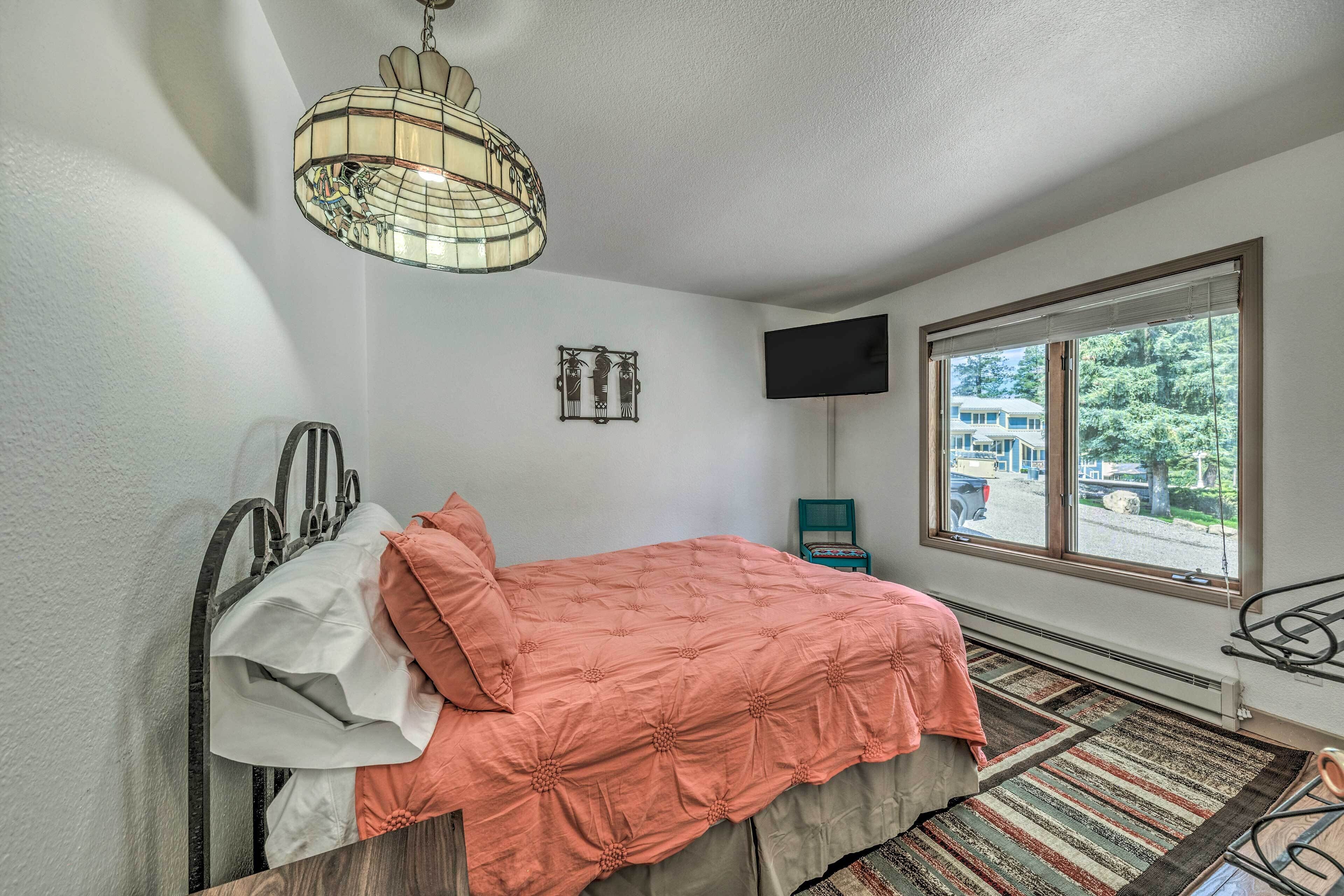 A bedroom with a bed, a hanging light fixture, a TV, and a window with a view of trees and a building.