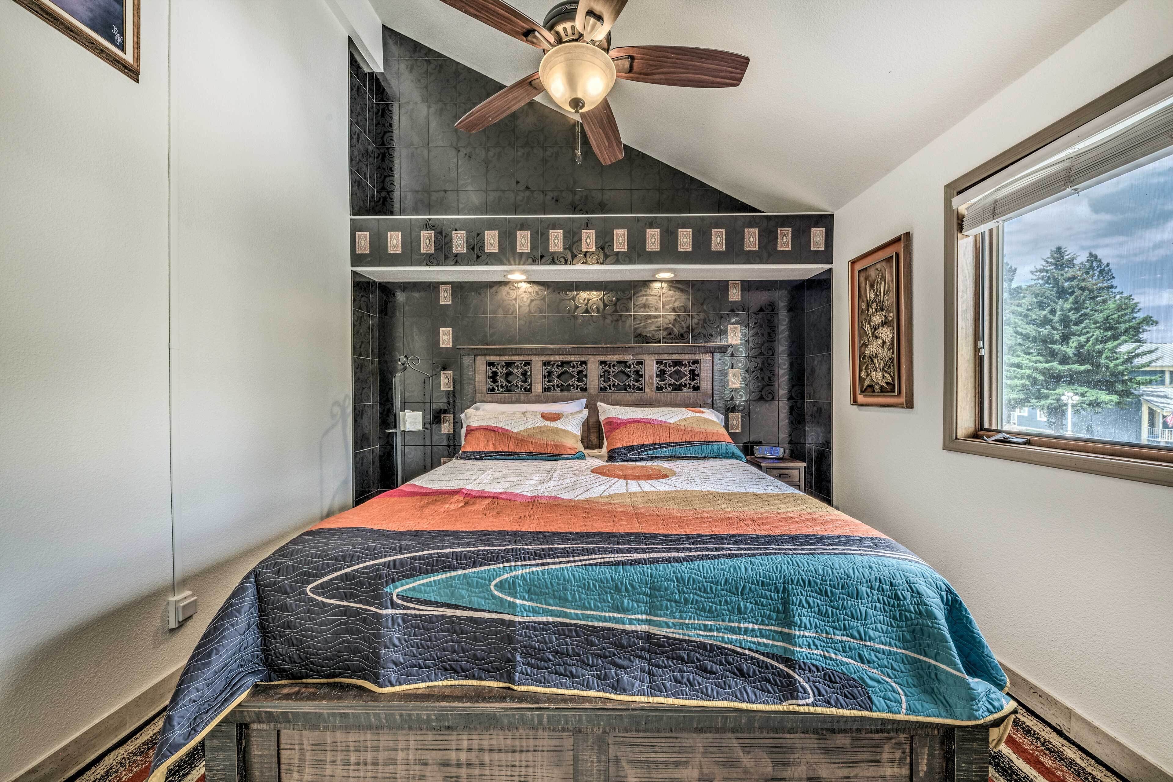 Un dormitorio con una cama grande, un ventilador de techo, una ventana con vista a árboles y un panel decorativo en la pared.