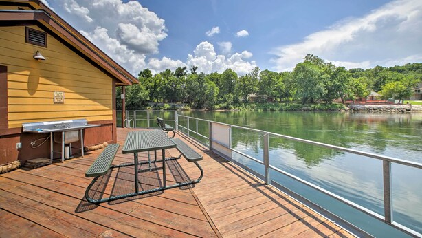 A wooden deck with picnic tables and a grill, overlooking a calm lake with a wooden dock and a yellow building with a sign.