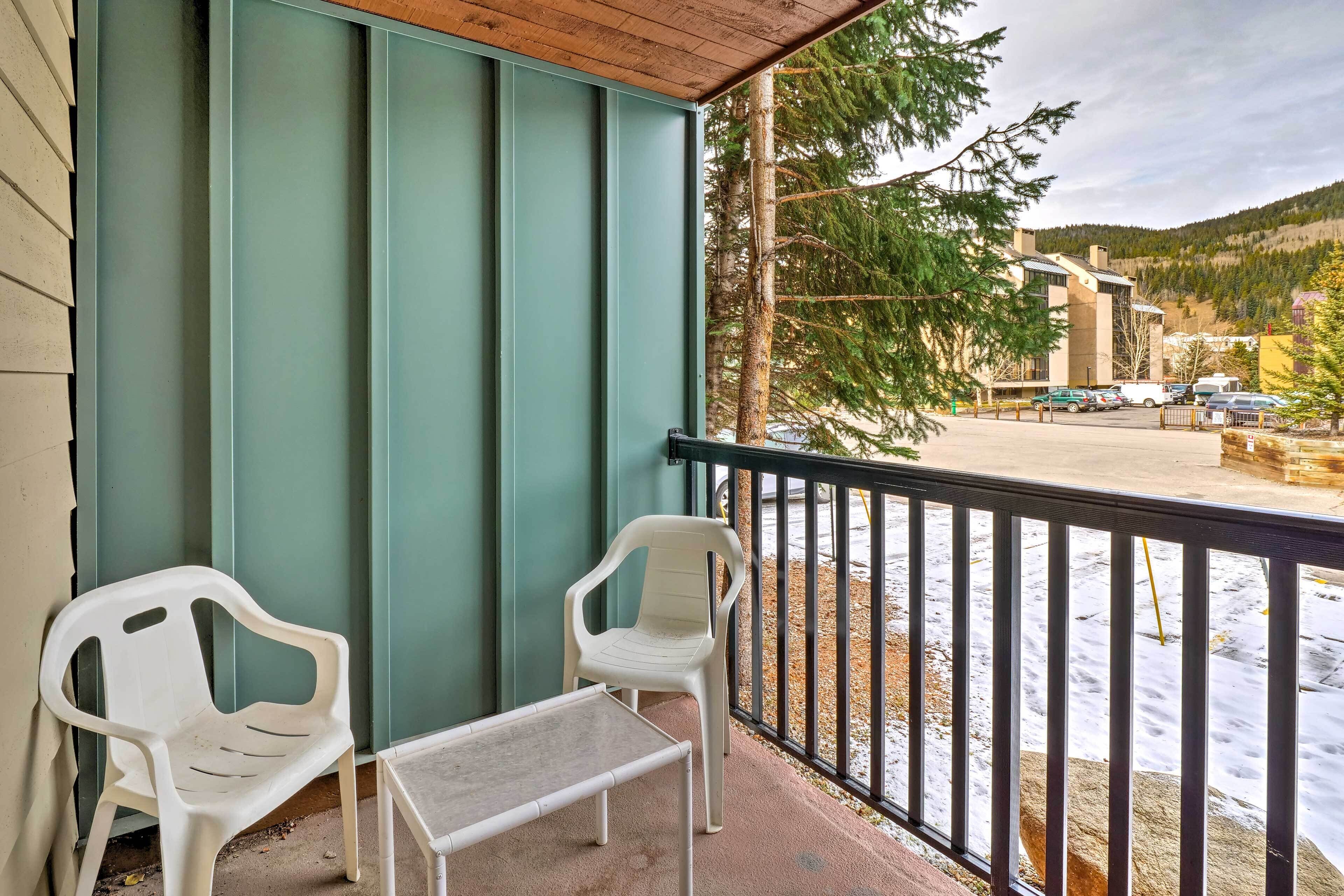 A balcony with two white plastic chairs and a small table, overlooking a parking area and a building with a green facade.