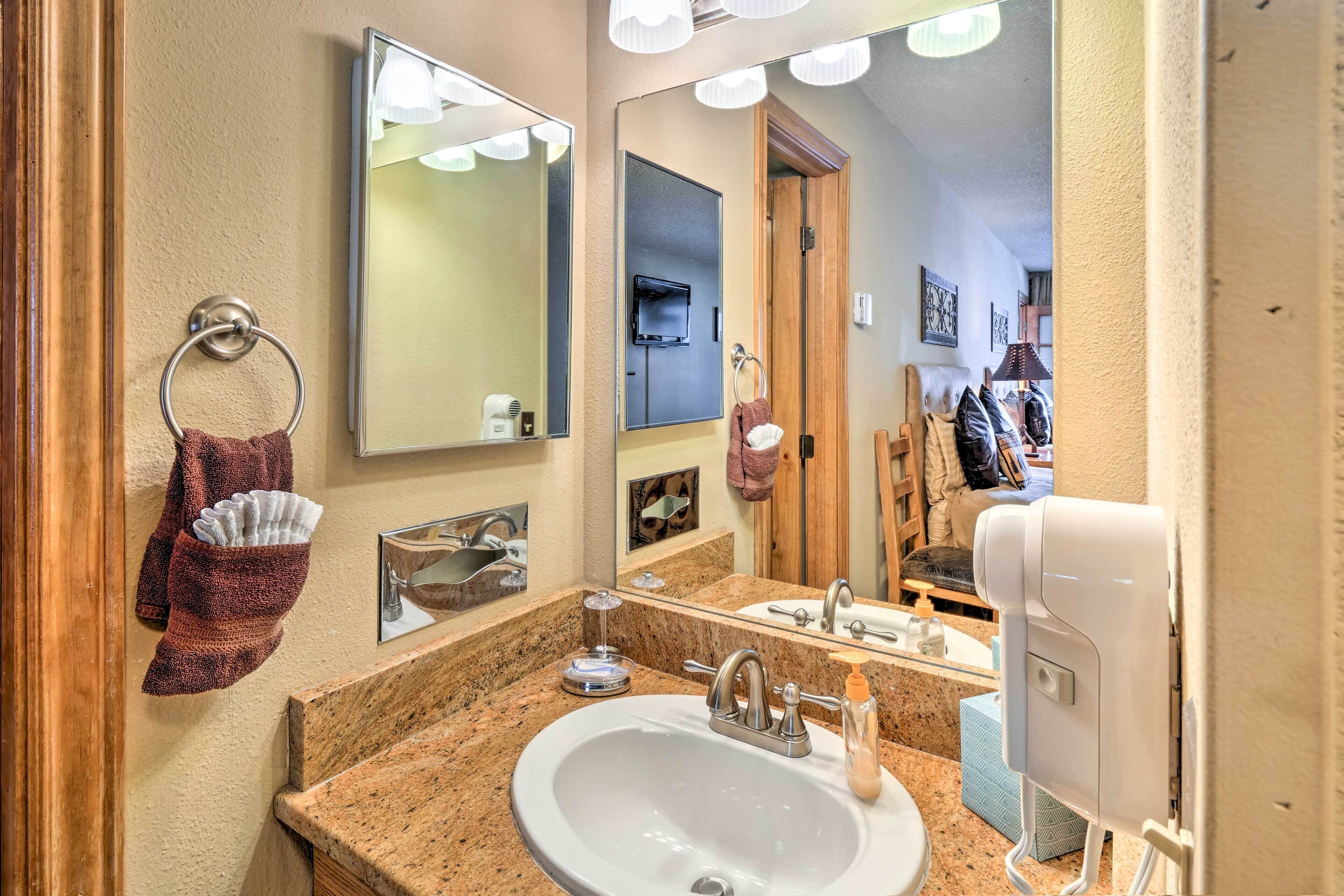 A bathroom with a granite countertop sink, a large mirror, a towel ring with a towel, a soap dispenser, and a handheld showerhead.
