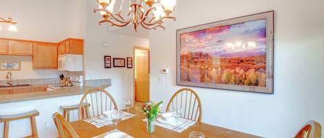 A dining area with a wooden table set for four, surrounded by chairs, a chandelier, and a painting of a landscape on the wall.
