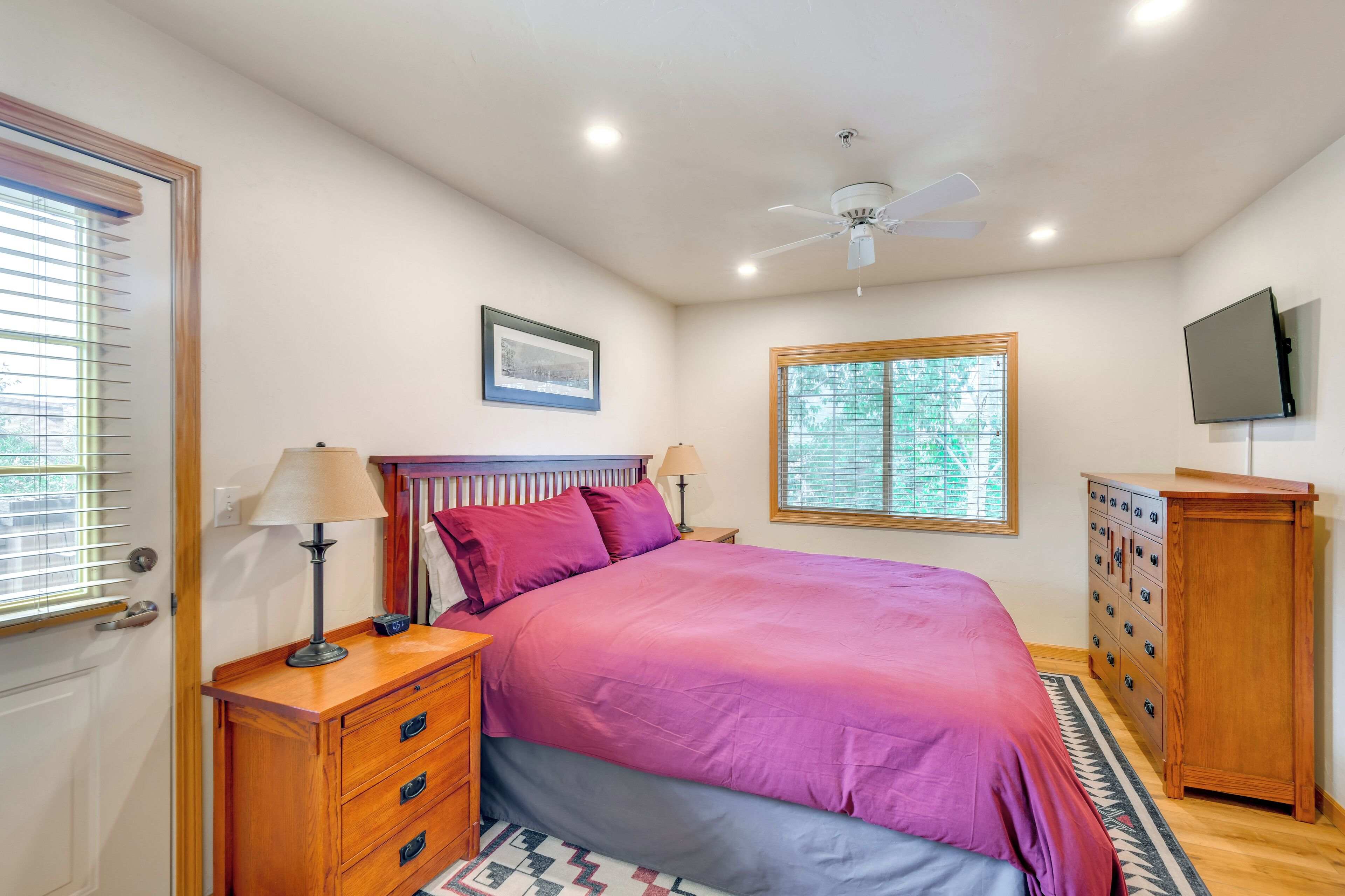 A bedroom with a bed, wooden dresser, nightstand, lamp, ceiling fan, window, and TV.