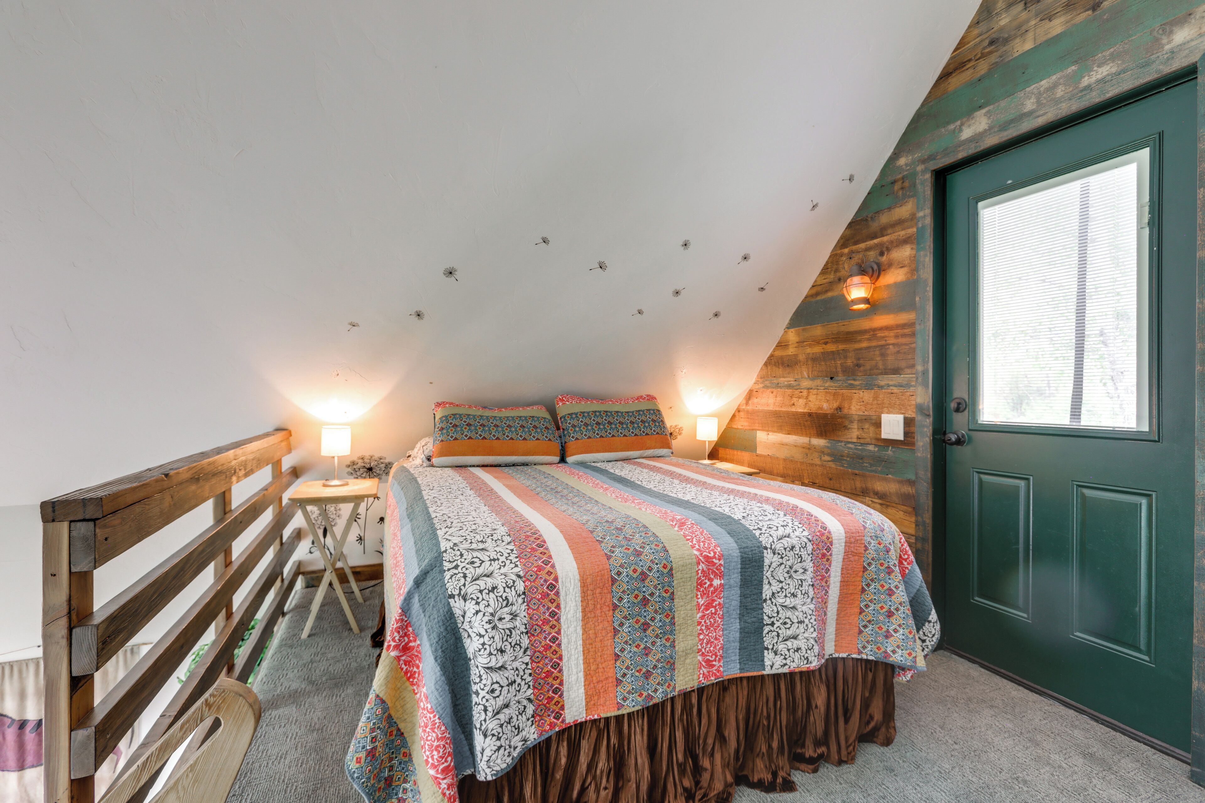 Une chambre avec un plafond en pente, une tête de lit en bois, un lit recouvert d’une courtepointe colorée, deux lampes de chevet et une porte verte.