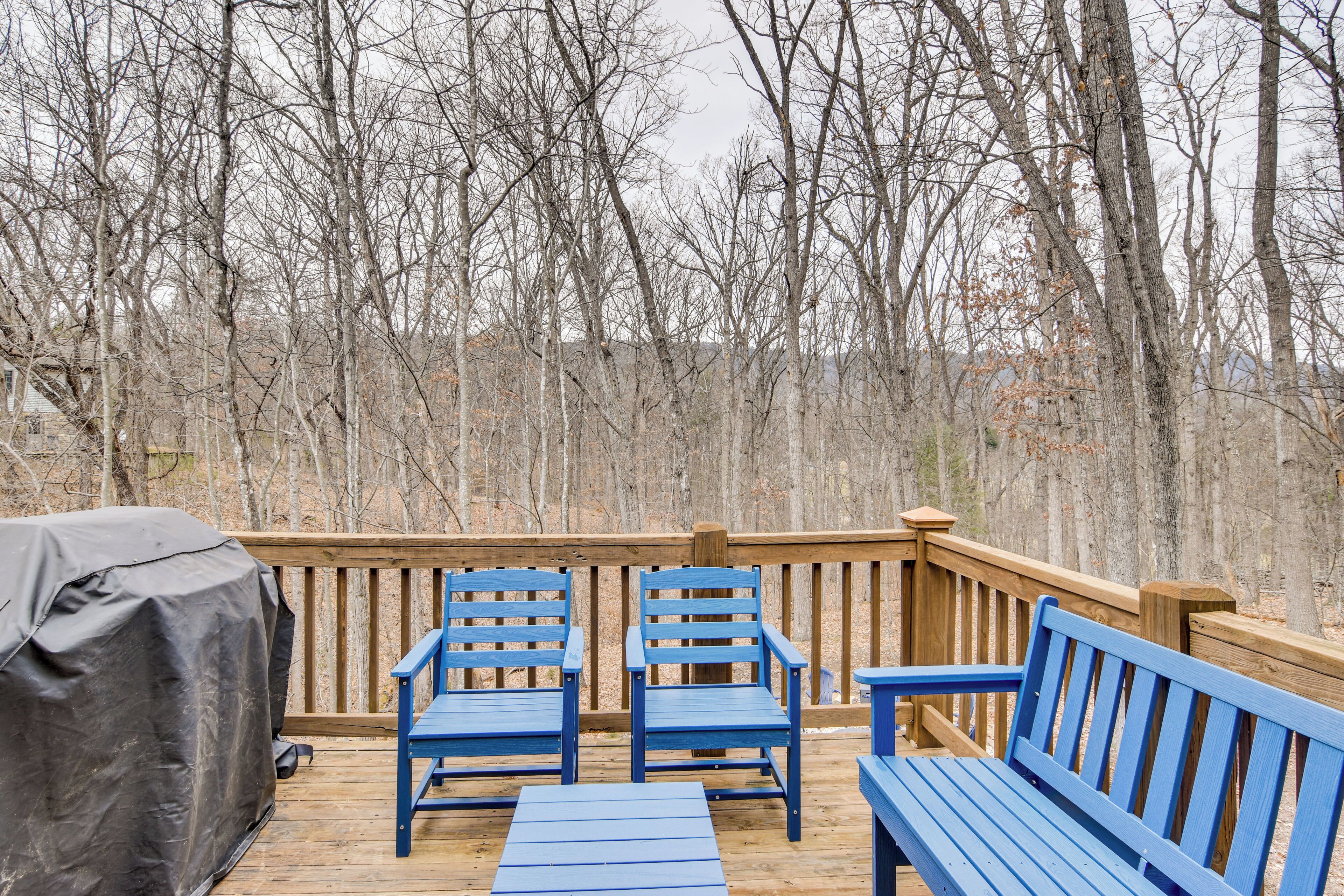 A wooden deck with blue outdoor furniture, including two chairs and a bench, surrounded by leafless trees.