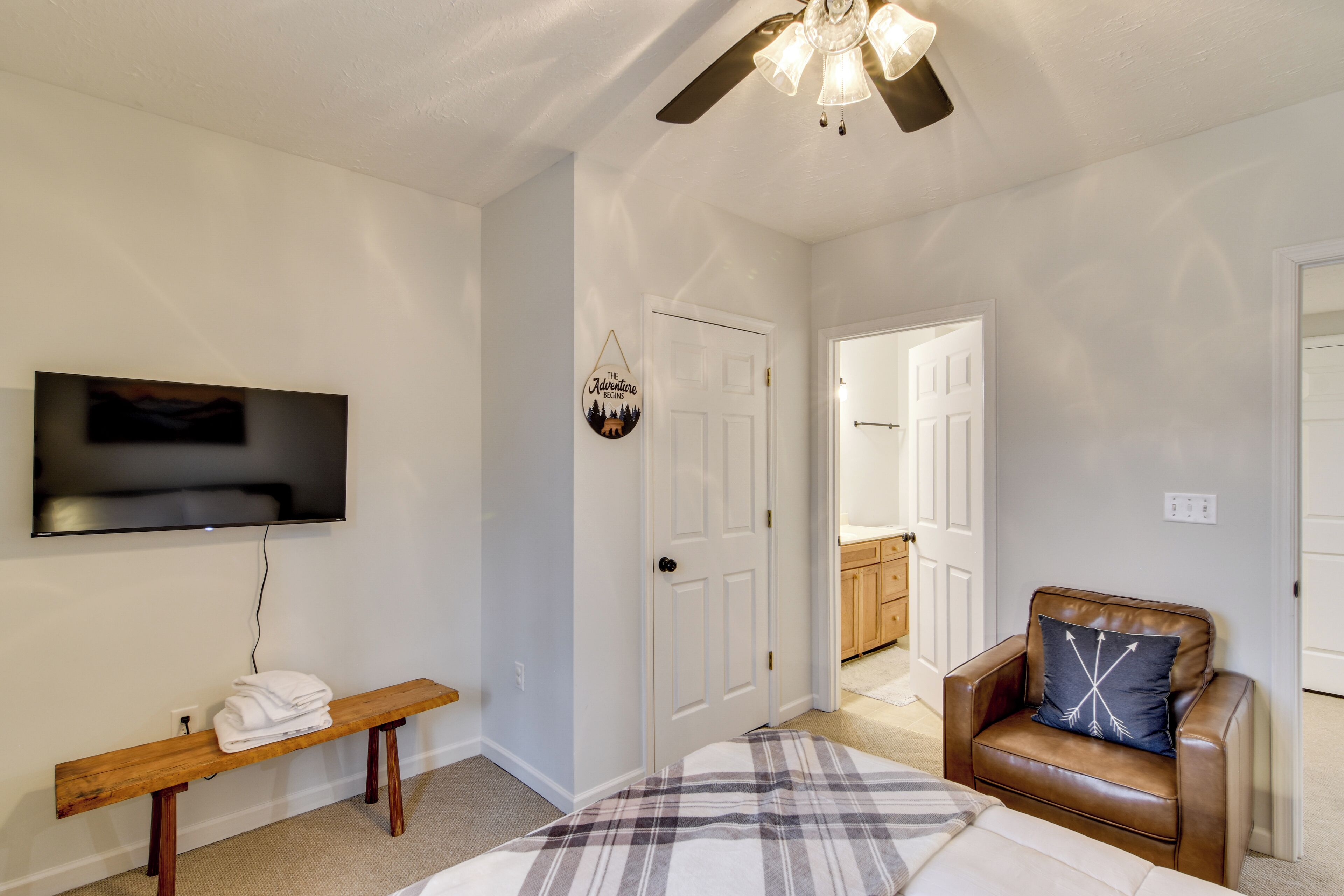 A bedroom with a bed, a bench, a chair, a ceiling fan, and a TV mounted on the wall.