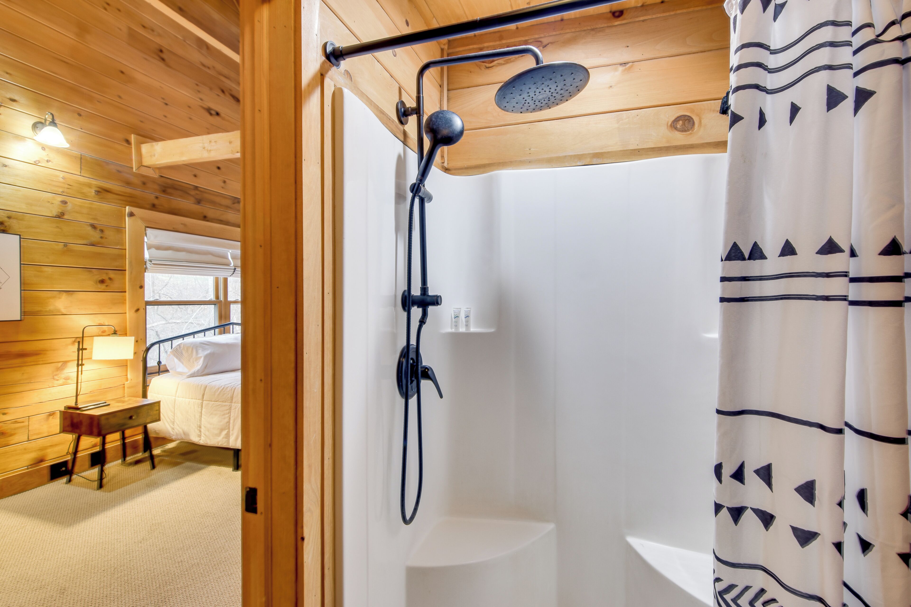 A wooden cabin interior with a shower, a bed, a small table, and a lamp.