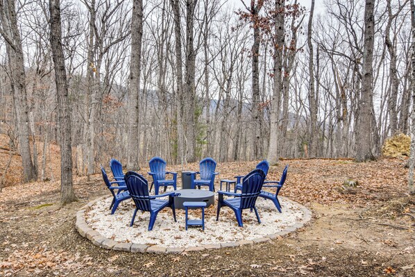 A circular outdoor seating area with blue chairs and a table surrounded by a stone border, set in a forested area with leaf-covered ground.
