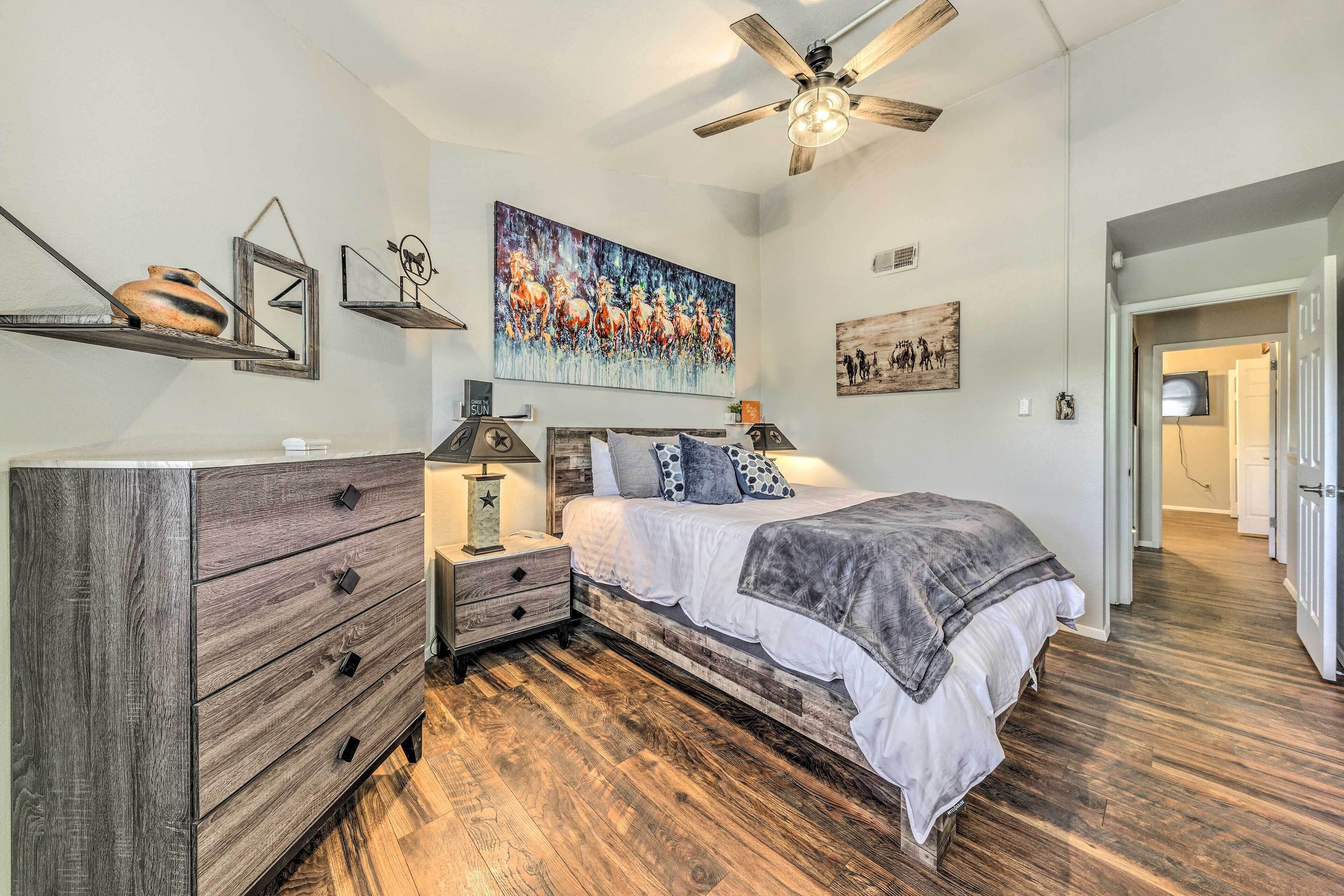 A bedroom with a wooden dresser, a bed with a gray blanket, a ceiling fan, and artwork on the walls.