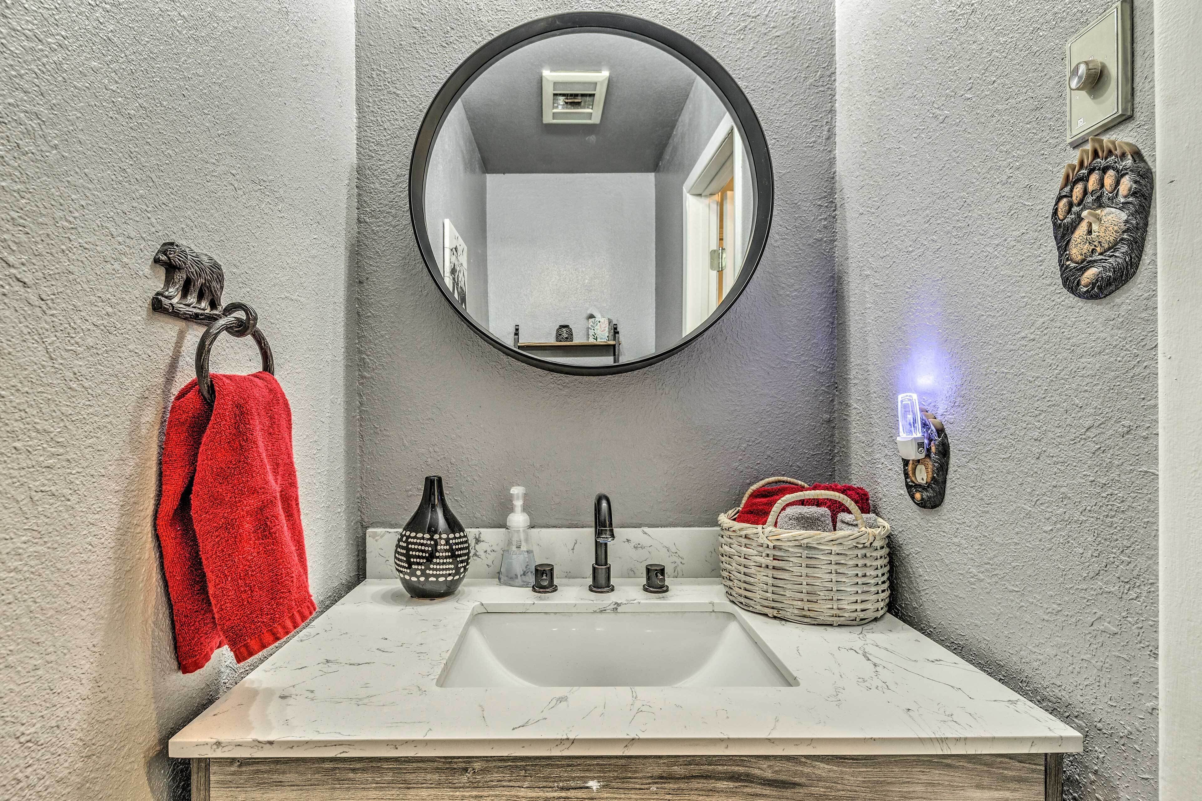 A bathroom with a round mirror, a white sink, a red towel, a black vase, a soap dispenser, and a basket.