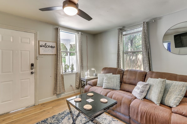 A living room with a brown leather sofa, a coffee table with decorative items, a ceiling fan, and a welcome sign on the door.
