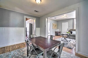 Un comedor con mesa de madera y sillas de metal, un tapete estampado y vista hacia una sala de estar con sofá y mesa de centro.