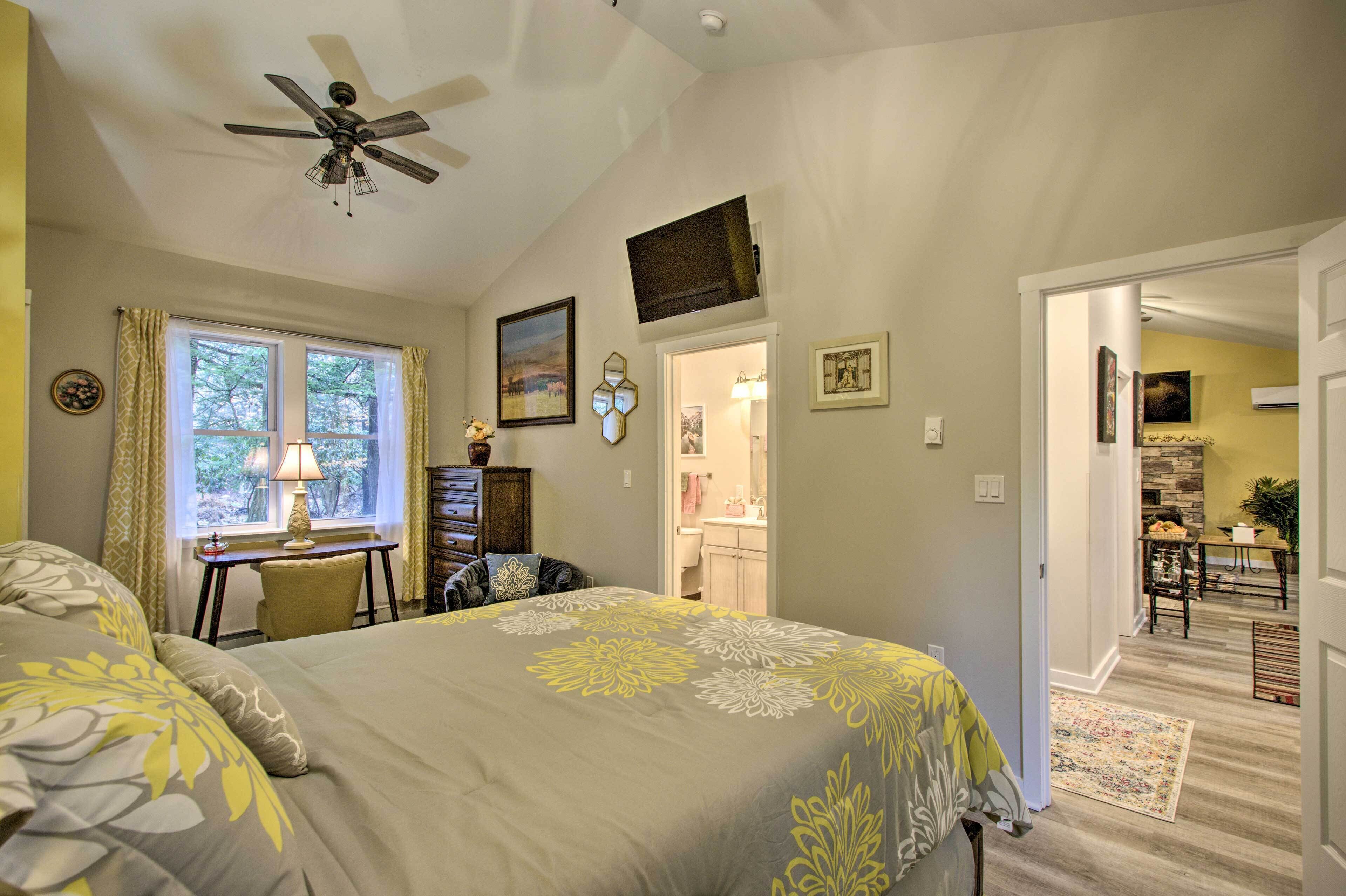 A bedroom with a bed, a desk, a chair, a ceiling fan, and a bathroom visible through an open door.