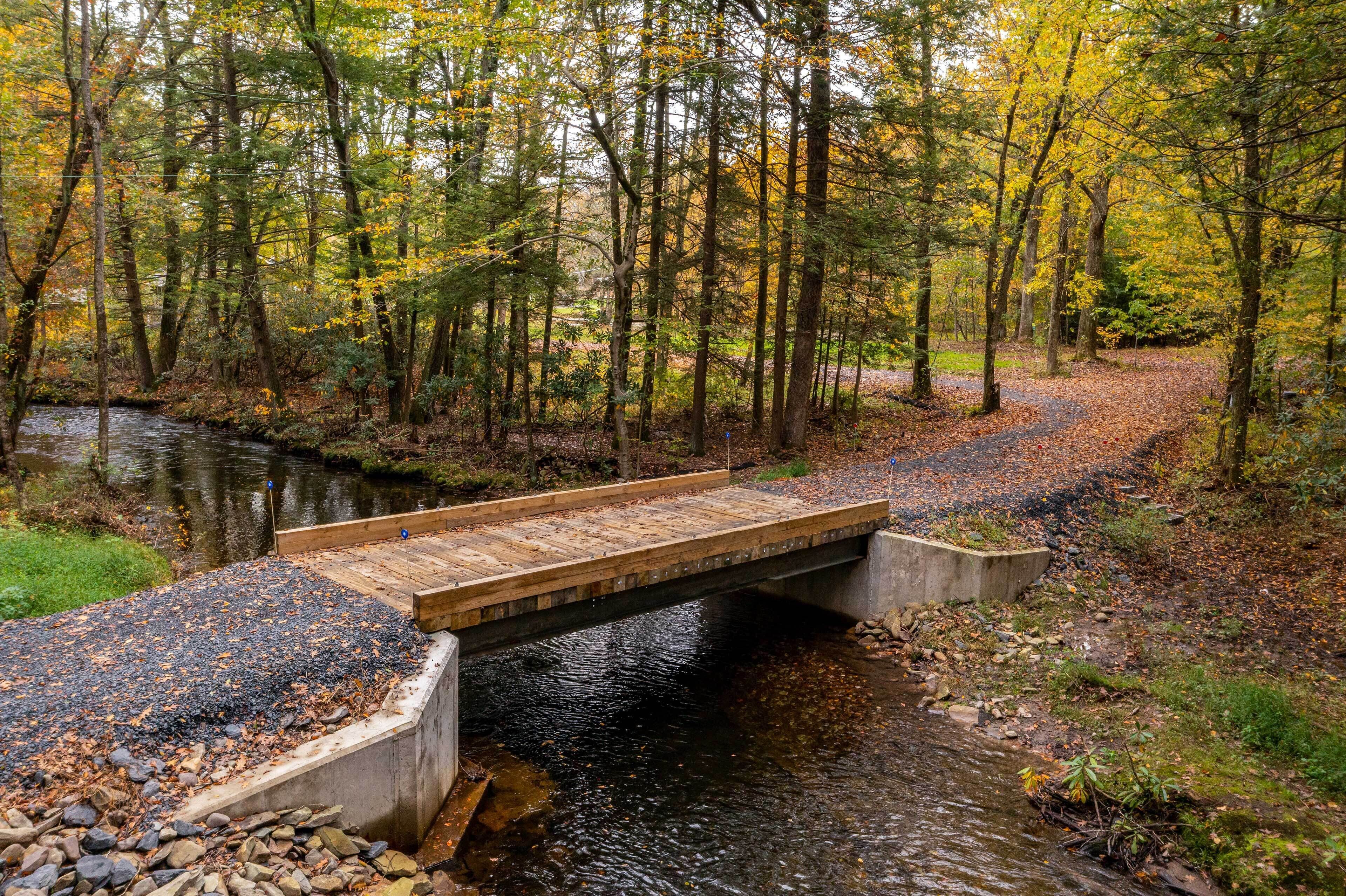 Un pont en bois enjambant un ruisseau dans une zone boisée, avec des feuilles d’automne.
