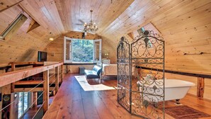 A wooden cabin interior with a wooden staircase, a bed, a bathtub, and a large window.
