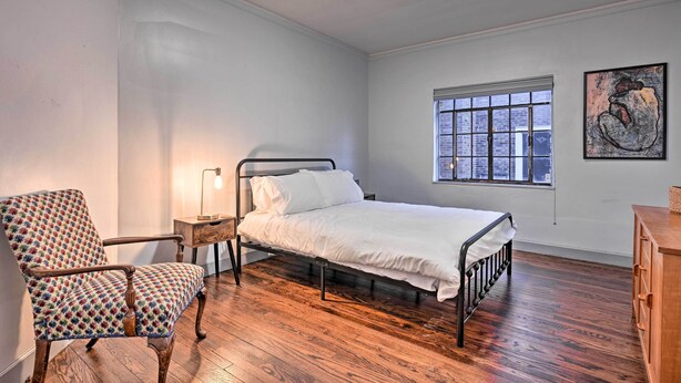 A bedroom with a bed, a chair, a nightstand, a lamp, a window, and a painting on the wall.