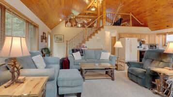 A spacious living area with a high wooden ceiling, a staircase leading to an upper level, and a kitchen area.