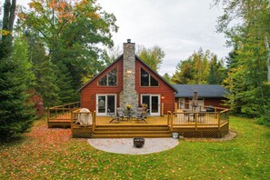 A cabin with a stone chimney, a deck with outdoor furniture, and a surrounding forest. - Elcho Escape w/ Private Dock on Post Lake! (Elcho)