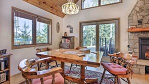 A wooden dining table with chairs, a chandelier, a stone fireplace, and large windows with a view of trees. - Elcho Escape w/ Private Dock on Post Lake! (Elcho)