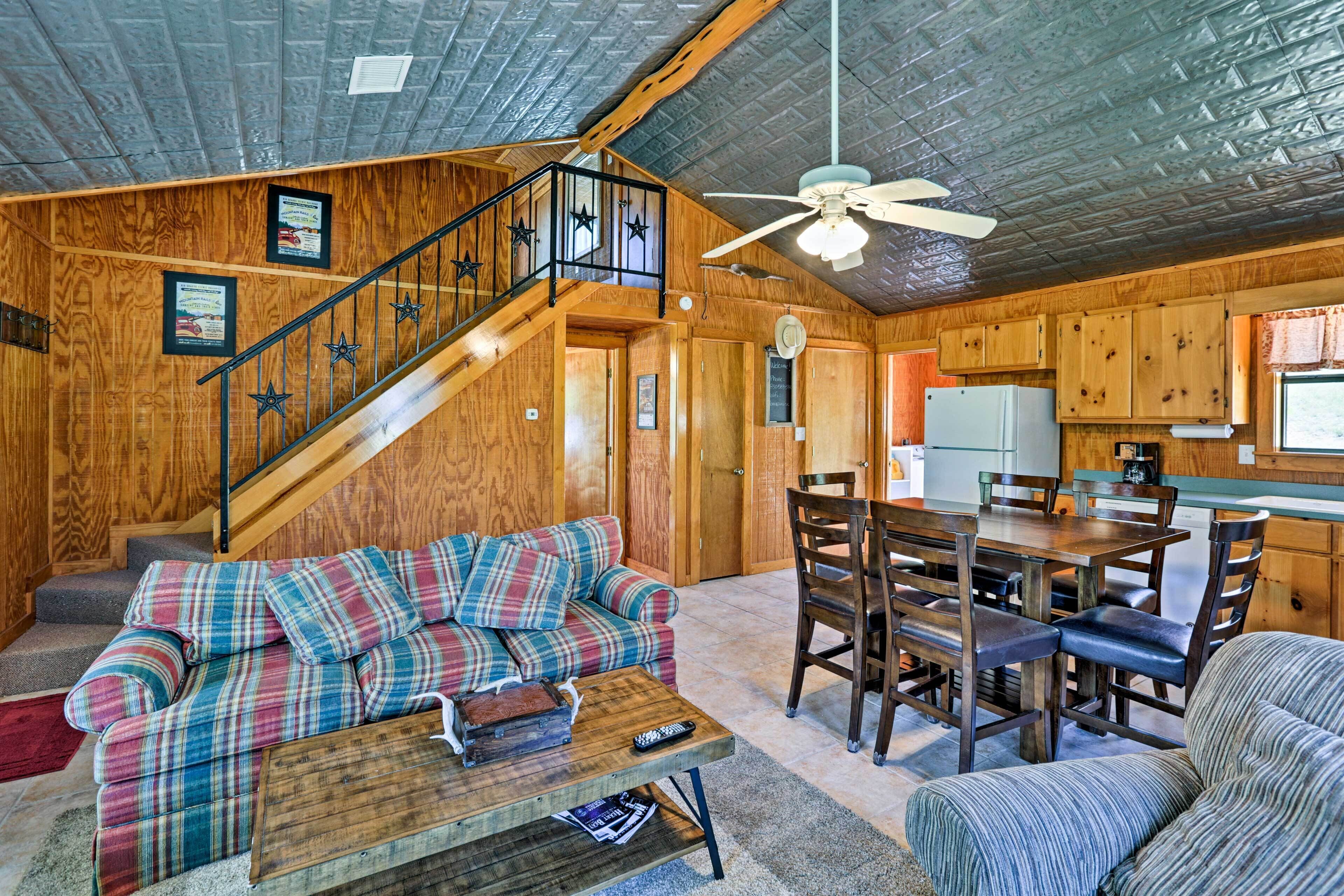 Interior acogedor de una cabaña con escalera de madera, sofá a cuadros, mesa de comedor y zona de cocina.