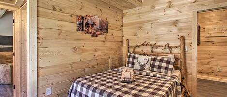 Une chambre à coucher dans une cabane en bois, avec une couvre-lit à carreaux, une tête de cerf fixée au-dessus de la tête de lit et un tableau représentant des cerfs accroché au mur.