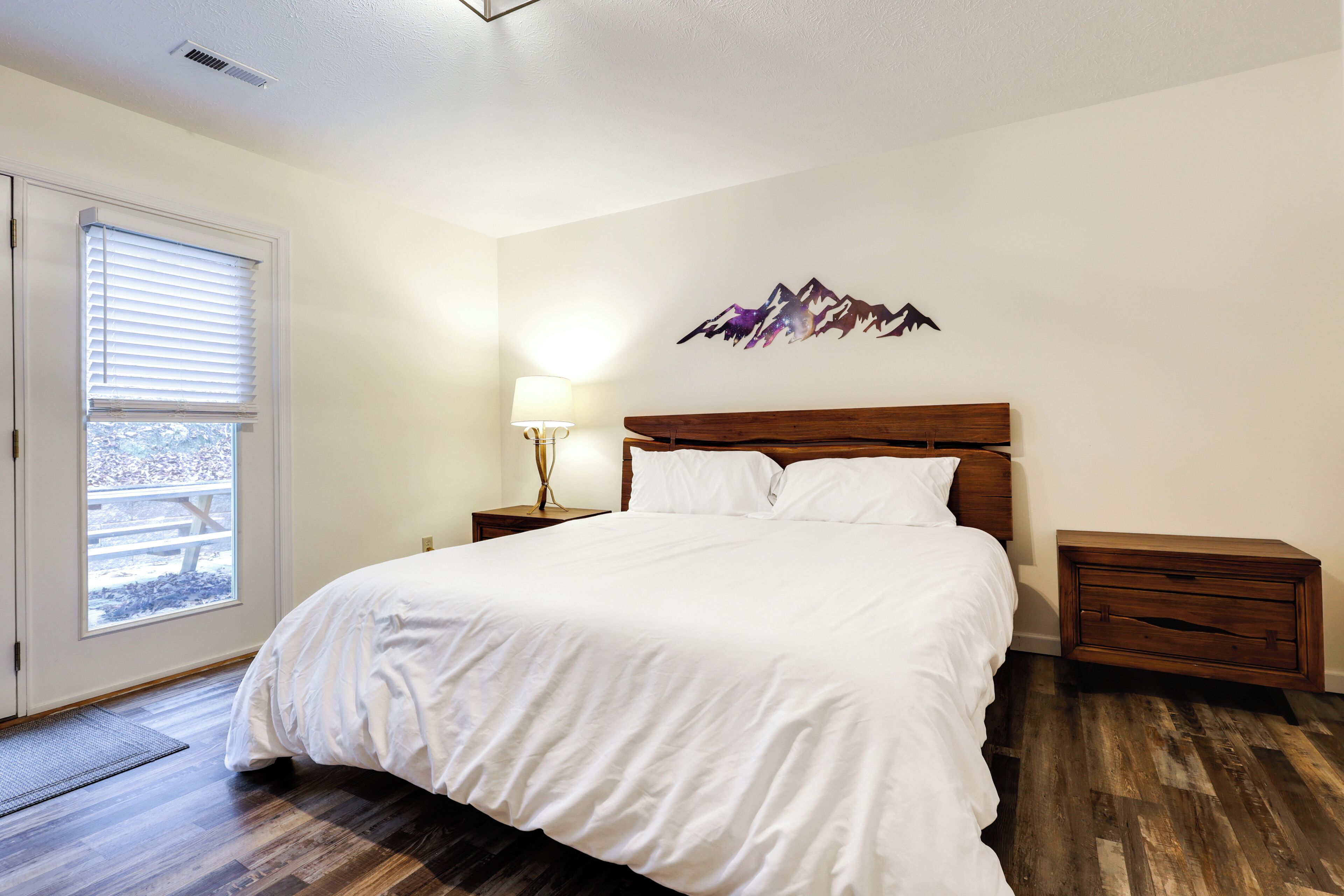 A bedroom with a large bed, a nightstand, a lamp, and a wall art piece.