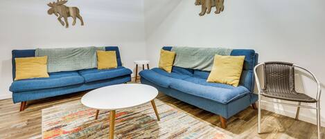 Una sala de estar con dos sofás azules, una mesa de centro blanca, un tapete estampado y una silla de mimbre.