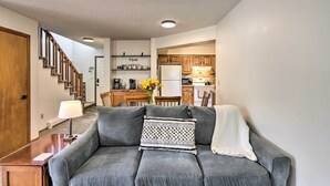 Un salon avec un canapé gris, une table basse en bois et une cuisine en arrière-plan.