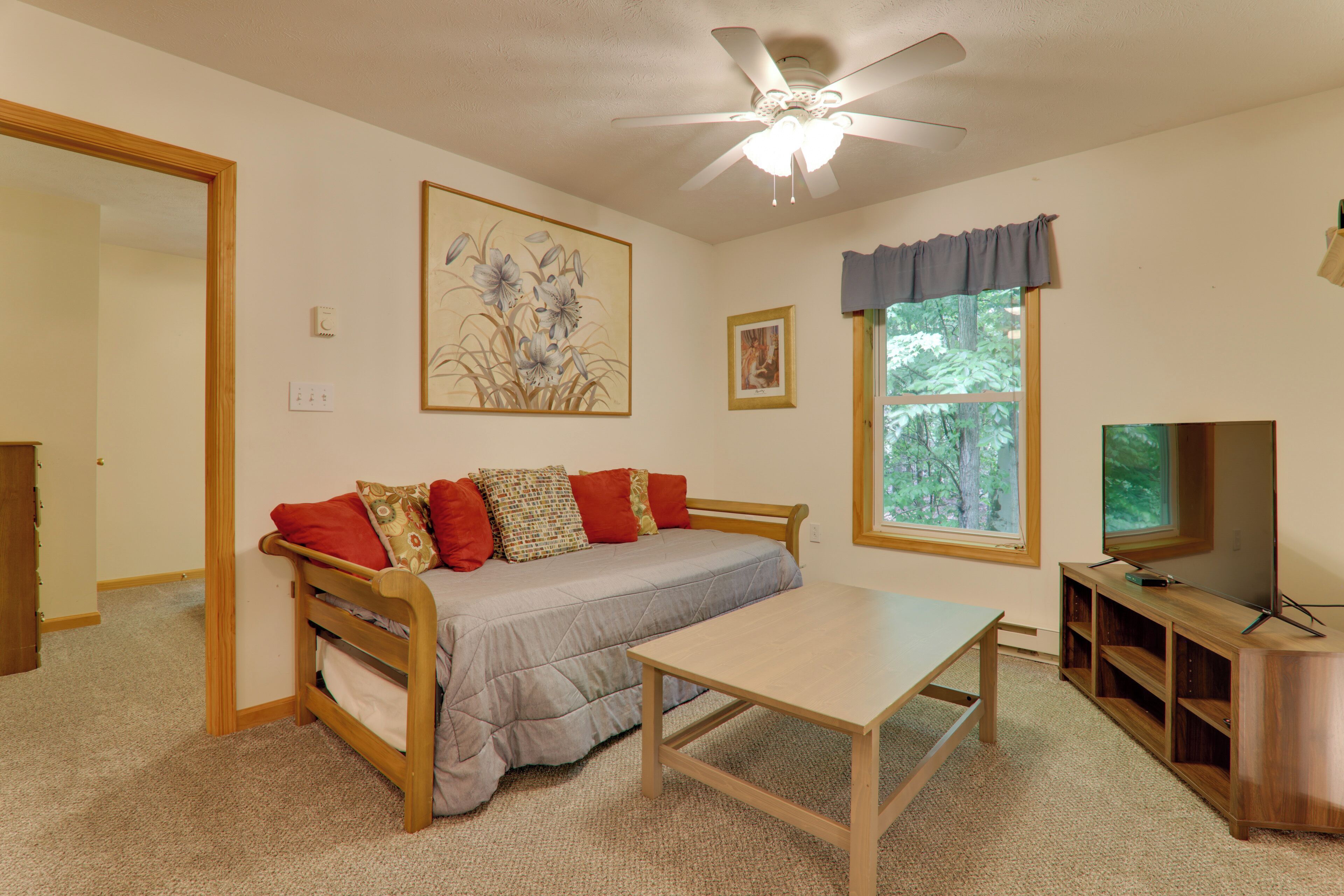 A bedroom with a bed, a table, a TV, a ceiling fan, and a window with a view of trees.