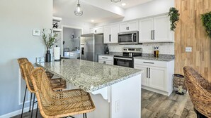 A modern kitchen with white cabinets, a granite countertop, and stainless steel appliances. There are wicker chairs and pendant lights. - Reeds Spring Condo ~ 3 Mi to Silver Dollar City! (Branson West)